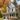 A picturesque two-story light blue Victorian-style house stands gracefully under a bright blue sky with white clouds, surrounded by vibrant autumn trees displaying rich orange, yellow, and red foliage. A white wrap-around porch with ornate columns and railings leads to a dark wood front door, with a curving gray gravel path and lush green lawn in the foreground adorned with pink flowering shrubs.