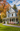A picturesque two-story light blue Victorian-style house stands gracefully under a bright blue sky with white clouds, surrounded by vibrant autumn trees displaying rich orange, yellow, and red foliage. A white wrap-around porch with ornate columns and railings leads to a dark wood front door, with a curving gray gravel path and lush green lawn in the foreground adorned with pink flowering shrubs.