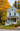 A charming two-story light blue clapboard house with white trim and a white wraparound porch stands prominently. The house is surrounded by vibrant golden-orange autumn trees and dark green pine trees, while the green lawn, gray sidewalk, and paved road below are scattered with fallen leaves. A potted plant with red flowers adds a touch of color to the porch, completing this inviting autumnal scene.