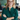A smiling young blonde woman with a warm expression looks directly at the camera. She wears round black-rimmed glasses and a dark green patterned blouse, with her arms crossed over her chest. The background is a softly blurred, modern office setting with warm, ambient lighting.