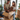 A young Caucasian couple smiles happily while unpacking cardboard boxes in their new home. The man, with a beard and light brown hair, reaches into an open box while the woman, with curly light brown hair, holds a small, fluffy brown dog. They are surrounded by numerous moving boxes and green houseplants, with large, bright windows in the background, conveying a sense of warmth and excitement for their fresh start.