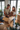 A young Caucasian couple smiles happily while unpacking cardboard boxes in their new home. The man, with a beard and light brown hair, reaches into an open box while the woman, with curly light brown hair, holds a small, fluffy brown dog. They are surrounded by numerous moving boxes and green houseplants, with large, bright windows in the background, conveying a sense of warmth and excitement for their fresh start.