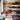A vertical shot showing numerous decorative pillows of various colors, patterns, and textures stacked on white rustic shelves and on the floor in an indoor setting. The pillows feature a mix of floral, geometric, and animal patterns in rich reds, yellows, creams, blacks, and blues, creating a vibrant, eclectic display. A wooden beam is visible on the far left, adding to the rustic charm.