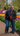 A happy elderly couple, a woman with short brown hair and a man with a grey beard, stand smiling together in a colorful tulip garden. They are both wearing dark jackets, blue jeans, and vibrant scarves, with rows of red, yellow, orange, purple, and white tulips blooming brightly behind and around them on a brown pathway.