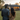 A full-length, back-view shot of a young couple standing on a manicured green lawn at golden hour. They are looking towards a modern, single-story house in the background, with warm light glowing from its windows against a soft, peachy sunset sky. The man on the left wears a dark gray shirt, and the woman on the right, with long dark hair, wears a camel-colored sweater, with her arm linked with his, conveying intimacy and shared focus.