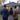View from behind three people standing on a green lawn, looking towards a suburban house at dusk. A man in a dark blue suit stands on the left, a woman in a patterned dress is in the middle, and a man in a brown suit is on the right. The gray house in the background has a dark roof, warm glowing windows, and the house number 823 is visible on the siding.