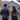 A rear view of a man and a woman standing on a neatly mowed green lawn, looking towards a light blue-grey suburban house. The man wears a dark suit jacket, and the woman wears a mustard-colored jacket with long dark hair. The house in the background features a dark roof, visible windows with interior light, and a glowing warm front door, conveying a sense of welcome and potential home ownership.