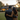 A diverse couple is viewed from behind, embracing and looking at a large, modern house in the distance. The man has dark, neatly styled hair and a beard, wearing a dark charcoal blazer over a light shirt, while the woman has long, flowing dark hair and wears a light brown or camel-colored sweater. They stand on a vibrant green lawn under a soft, golden sunset sky, with the house's windows illuminated warmly, symbolizing hope, aspiration, and the comfort of home.