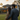 A man and a woman, viewed from behind, stand in a verdant green lawn, looking towards a large, modern house at sunset. The man, with dark hair and a beard, has his arm around the woman, whose long dark hair is illuminated by the warm, golden light of the setting sun. The two-story house in the softly blurred background features numerous windows glowing with interior lights, a dark grey roof, and a chimney, all set against a soft, golden-orange sky with faint clouds.