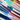A close-up, high-angle shot of numerous thick, glossy paint swatches arranged diagonally in parallel lines on a pure white surface. The swatches display a wide range of vibrant colors, including various shades of blue, green, brown, purple, pink, and red, with visible brushstroke textures that suggest thick applications of acrylic or oil paint. The diagonal orientation of the vibrant, overlapping colors creates a dynamic, colorful striped pattern from the bottom left to the top right of the frame.
