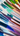 A close-up, high-angle shot of numerous thick, glossy paint swatches arranged diagonally in parallel lines on a pure white surface. The swatches display a wide range of vibrant colors, including various shades of blue, green, brown, purple, pink, and red, with visible brushstroke textures that suggest thick applications of acrylic or oil paint. The diagonal orientation of the vibrant, overlapping colors creates a dynamic, colorful striped pattern from the bottom left to the top right of the frame.
