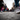 A close-up, low-angle view of a person's feet wearing grey patterned sneakers and blue jeans, walking forward on a dark grey asphalt street. A distinct white line runs across the pavement, and a large, blurred crowd of people in various colorful attire walks in the background, conveying dynamic motion in an urban setting.