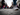 A close-up, low-angle view of a person's feet wearing grey patterned sneakers and blue jeans, walking forward on a dark grey asphalt street. A distinct white line runs across the pavement, and a large, blurred crowd of people in various colorful attire walks in the background, conveying dynamic motion in an urban setting.