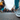 A low-angle, eye-level photograph of a person's feet wearing grey sneakers and blue jeans, sharply in focus as they walk on a textured grey asphalt street. Behind them, a large crowd of people is blurred with motion, suggesting a busy urban environment, with indistinct city buildings lining the street under an overcast sky. The image conveys the dynamic energy and movement of city life.