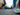 A low-angle, eye-level photograph of a person's feet wearing grey sneakers and blue jeans, sharply in focus as they walk on a textured grey asphalt street. Behind them, a large crowd of people is blurred with motion, suggesting a busy urban environment, with indistinct city buildings lining the street under an overcast sky. The image conveys the dynamic energy and movement of city life.