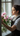 A young woman with dark hair pulled back is shown in profile, looking out a window as she carefully arranges a bouquet of pink peonies. She wears a white collared shirt and a dark apron, with her face and the flowers softly illuminated by natural light streaming through the multi-pane window. Her hands hold a clear glass vase filled with vibrant pink peony blooms and green foliage, positioned on a light-colored windowsill.