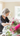 A mature woman with short, stylish gray hair, wearing a dark gray long-sleeved shirt and a subtle silver necklace, is seen in a close-up, arranging a bouquet of pink and fuchsia flowers. She is looking down at the flowers with a gentle smile, her hands carefully placing stems into the arrangement. The scene is bathed in soft natural light coming from a window in a warm-toned room, with some blurred pink flowers in the foreground.