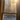 A vertical image looking out through a traditional wooden-framed window. The window glass is covered in a shimmering layer of frost or ice, catching the light. Golden sunlight streams intensely from the upper right corner, creating a brilliant starburst effect and brightly illuminating the ice particles on the glass. Through the frosted panes, bare trees and a snow-covered ground under a clear blue sky are visible in the distance, with a small snowdrift resting on the lower window sill.