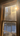 A vertical image looking out through a traditional wooden-framed window. The window glass is covered in a shimmering layer of frost or ice, catching the light. Golden sunlight streams intensely from the upper right corner, creating a brilliant starburst effect and brightly illuminating the ice particles on the glass. Through the frosted panes, bare trees and a snow-covered ground under a clear blue sky are visible in the distance, with a small snowdrift resting on the lower window sill.
