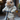 A light brown, fluffy rabbit stands upright on a miniature black kick scooter. It is wearing a cream-colored knitted sweater and matching pants with small colorful flecks, holding onto the scooter's handlebars. The background is softly blurred, showing muted tones of an outdoor or urban setting.