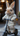 A light brown, fluffy rabbit stands upright on a miniature black kick scooter. It is wearing a cream-colored knitted sweater and matching pants with small colorful flecks, holding onto the scooter's handlebars. The background is softly blurred, showing muted tones of an outdoor or urban setting.