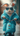 A full-body vertical shot of a white rabbit with fluffy fur, wearing a bright turquoise furry zippered hoodie and reflective silver-framed sunglasses, riding a matching turquoise scooter. The rabbit's front paws are on the chrome handlebars, and its face is partially obscured by the dark mirrored lenses. The background is softly blurred with muted natural tones, suggesting an outdoor setting, creating a whimsical and stylish scene.
