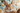 A very close-up, head-on shot of a fluffy brown rabbit with long whiskers and large, dark eyes looking directly forward. Its pink nose and slightly open mouth are prominent. Two blurry, pastel-colored eggs, one yellowish-orange and one peach, appear to float on either side of its head against a soft, light blue and white bokeh background. The rabbit's forepaws are visible at the bottom, suggesting a 'selfie' perspective.